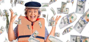 Gri saçlı, deniz cankurtaranı ceketi giyen mutlu bir gülümsemeyle zaferi kutlayan ve elleri havada kazanan bir ifade takınan yaşlı kadın.