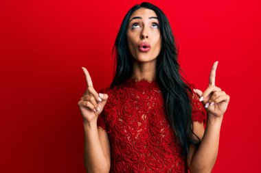 Kırmızı arka planda zarif elbiseler giyen güzel İspanyol kadın şaşırmış ve şaşırmış bir şekilde yukarı baktı ve parmaklarıyla işaret ederek kollarını kaldırdı.. 
