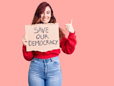 Elinde demokrasi protesto pankartımızı tutan genç ve güzel bir kadın mutlu ve pozitif gülümseyen, başparmağıyla mükemmel ve onay işareti yapan. 