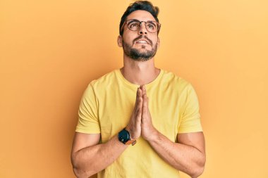 Gündelik kıyafetler ve gözlük takan genç İspanyol adam elleri ile yalvarıyor ve dua ediyor. Yüzünde umut ifadesi var. Çok duygusal ve endişeli. Yalvarıyor. 