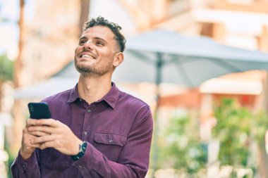 Genç İspanyol adam şehirde akıllı telefon kullanarak mutlu gülümsüyor..