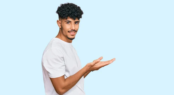 若いアラブ人男性を着てカジュアルな白いTシャツを指摘脇に手を開いてヤシの木を示すコピースペース 提示広告笑顔興奮幸せ — ストック写真