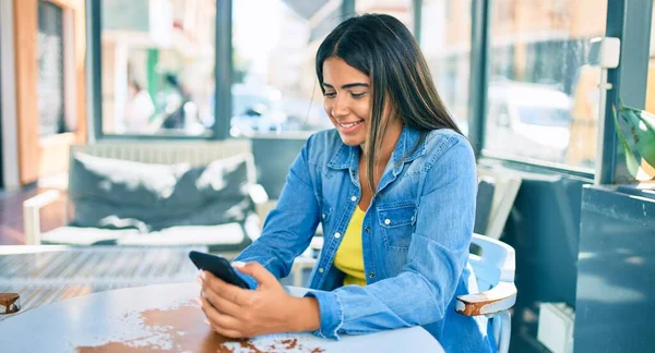 Young latin woman smiling happy using smartphone at coffee shop terrace.