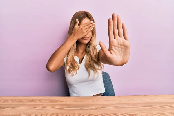 Beautiful blonde young woman wearing casual clothes sitting on the table covering eyes with hands and doing stop gesture with sad and fear expression. embarrassed and negative concept.