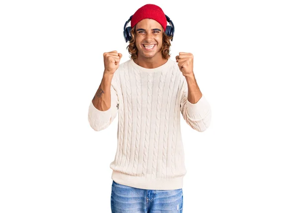 ヘッドフォンを使用して音楽を聴いている若いヒスパニック系の男性は 腕を上げて目を開いて成功したことに驚きと驚きを祝います 勝者のコンセプト — ストック写真