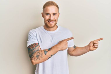 Beyaz bir tişört giymiş genç beyaz bir adam gülümsüyor ve kameraya bakıyor iki eliyle ve parmaklarıyla kenarı işaret ediyor.. 