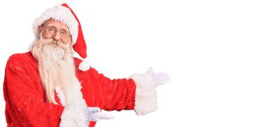 Old senior man with grey hair and long beard wearing traditional santa claus costume inviting to enter smiling natural with open hand 