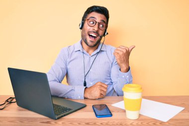 Çağrı merkezindeki operatör kulaklığı takmış, gülümseyen, gülümseyen ve baş parmağıyla yan tarafı gösteren İspanyol genç adam.. 