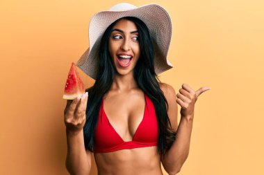 Güzel İspanyol kadın bikini giyip karpuz yiyor başparmağıyla yan tarafa bakıyor ve ağzı açık gülümsüyor. 