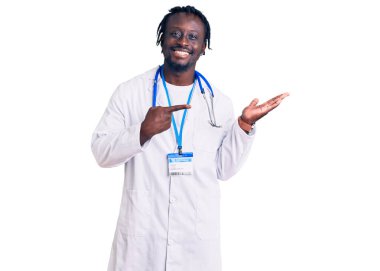 Doktor steteskop ve kimlik kartı takan genç Afrikalı Amerikalı adam kameraya hayret içinde bakıp gülümserken eliyle ve parmağıyla işaret ediyor.. 