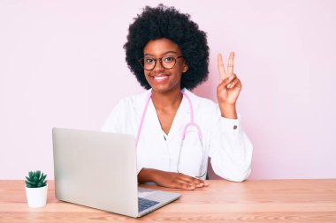 Doktor steteskobu takan Afro-Amerikalı genç bir kadın kameraya gülümseyen gülen bir bilgisayarla çalışıyor zafer işareti yapıyor. İki numara.. 