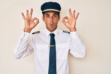 Uçak pilotu üniforması giyen genç İspanyol adam şaşırmış ve şaşırmış görünüyor. Tamam sembolünü parmaklarıyla yapıyor. çılgın ifade 