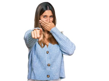 Gündelik kıyafetler giyen genç bir kadın sana gülüyor, eli ağzında kamerayı işaret ediyor, yüz ifadesi utanç verici. 