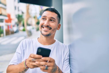 Duvara dayanmış akıllı telefon kullanan, mutlu gülümseyen genç İspanyol adam.
