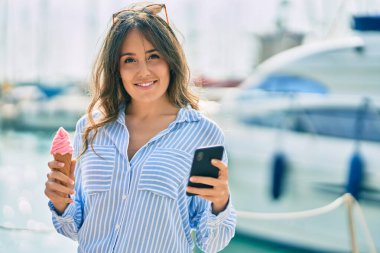 Genç İspanyol kadın akıllı telefon kullanıyor ve limanda dondurma yiyor..