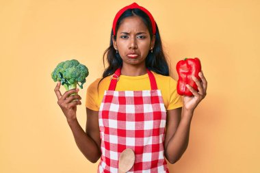 Young indian girl wearing apron holding broccoli and red pepper depressed and worry for distress, crying angry and afraid. sad expression. 