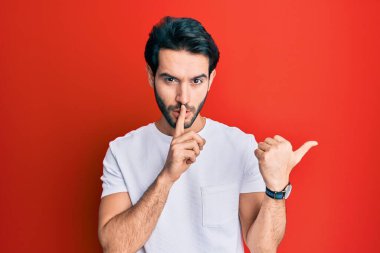 Gündelik beyaz tişört giyen genç İspanyol adam sessiz olmak istiyor. Parmakları dudaklarında ve elleri yandan işaret ediyor. sessizlik ve gizli konsept. 