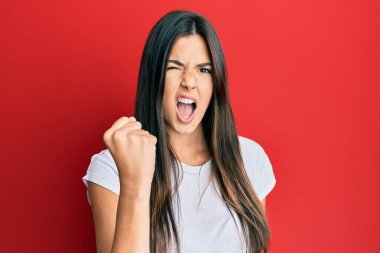 Genç esmer kadın, kırmızı arka plan üzerine gündelik beyaz tişört giyip öfkeyle bağırırken sinirli ve öfkeli bir şekilde yumruk sallıyor. öfke ve agresif konsept. 