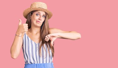 Young beautiful blonde woman wearing summer hat doing thumbs up and down, disagreement and agreement expression. crazy conflict 