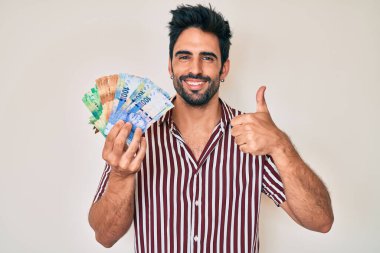 Sakallı yakışıklı İspanyol adam Güney Afrika rand banknotları tutuyor mutlu ve olumlu gülümsüyor, başparmağıyla mükemmel ve onay işareti yapıyor. 