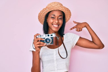 Young african american woman wearing summer hat holding vintage camera looking confident with smile on face, pointing oneself with fingers proud and happy. 