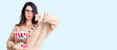 Beautiful young brunette woman eating popcorn with angry face, negative sign showing dislike with thumbs down, rejection concept 