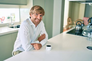 Genç İrlandalı adam gülümsüyor. Mutlu bir şekilde kahve içiyor. Evdeki masada oturuyor..