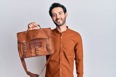 Deri çanta giyen genç İspanyol adam pozitif ve mutlu görünüyor. Dişlerini gösteren kendine güvenen bir gülümsemeyle ayakta duruyor ve gülümsüyor. 