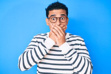 Gündelik süveter ve gözlük giyen genç, yakışıklı İspanyol adam yanlışlıkla elleriyle ağzını kapatırken şok oldu. gizli kavram. 