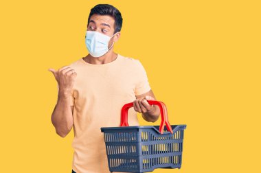 Alışveriş sepeti ve tıbbi maske takan genç İspanyol adam baş parmağını yukarı kaldırıyor ve ağzı açık gülümsüyor. 