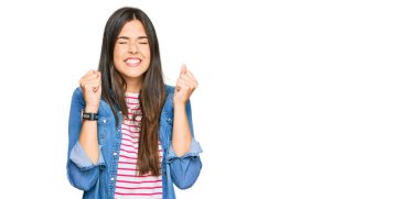 Gündelik kıyafetler giyen genç esmer kadın başarı için heyecanlı kolları havada ve gözleri kapalı zaferi kutlarken gülümsüyor. Kazanan konsept. 