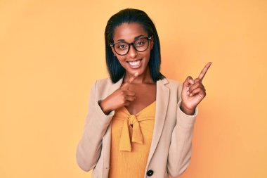 Genç Afro-Amerikan kadın iş kıyafetleri içinde gülümseyen ve kameraya bakıp iki eliyle ve parmaklarıyla kenarı işaret ediyor.. 