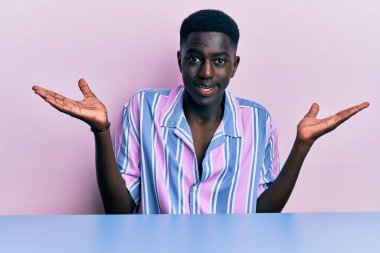 Gündelik giysiler giymiş, elleri ve kolları havada, kafası karışmış bir şekilde masada oturan Afro-Amerikalı genç bir adam. şüphe kavramı. 