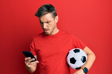 Futbol topu tutan yakışıklı beyaz adam akıllı telefona bakıyor kızgın ve kızgın bağırıyor sinirli ve öfkeli, öfkeyle bağırıyor. öfke ve agresif konsept. 