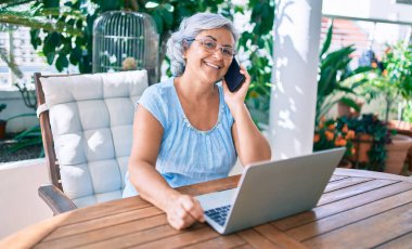 Gri saçlı, gülümseyen, mutlu, rahatlatıcı orta yaşlı bir kadın terasta oturuyor ve evden çalışıyor.