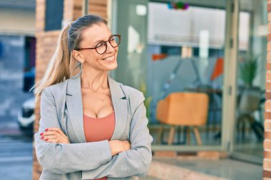 Sarışın iş kadını kollarını kavuşturup şehirde mutlu mesut gülümsüyor..