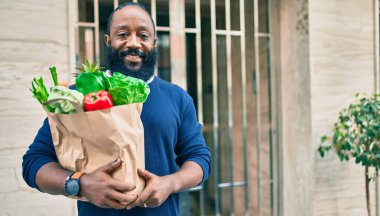 Sakallı Afrikalı Amerikalı bir adam elinde süpermarketten alınmış kağıt poşetler tutuyor.