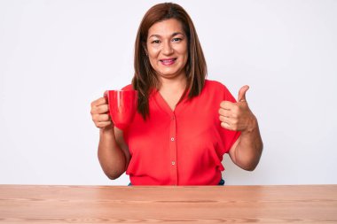 Orta yaşlı esmer kadın masaya oturmuş bir fincan kahve içiyor mutlu ve pozitif gülümsüyor, başparmağıyla mükemmel ve onay işareti yapıyor. 