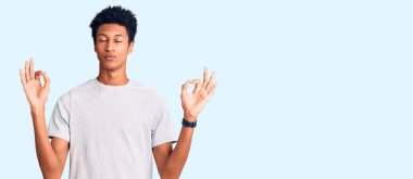 Genç Afro-Amerikan adam günlük kıyafetler giyip rahatlıyor ve gözleri kapalı gülümsüyor parmaklarıyla meditasyon hareketi yapıyor. yoga konsepti. 