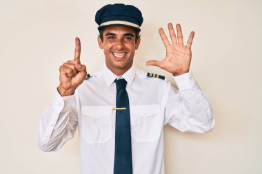 İspanyol asıllı genç adam uçak pilotu üniforması giyiyor. Altıncı parmaklarını gösterip işaret ederken kendinden emin ve mutlu gülümsüyor.. 