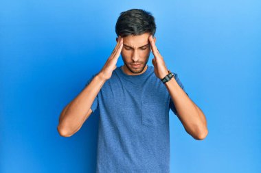 Mavi arka planda, elleri başının üstünde, stresten dolayı başı ağrıyan sıradan tişörtler giyen genç yakışıklı bir adam. migren ağrısı. 