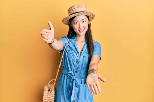 Young chinese woman wearing summer hat looking at the camera smiling with open arms for hug. cheerful expression embracing happiness.