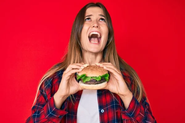 美しい白人女性はおいしい古典的なハンバーガーを食べて怒りと怒りの叫び不満と激怒怒りを叫び怒りを見上げ — ストック写真