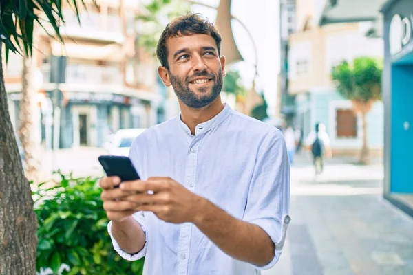 一个英俊的男人 留着胡子 穿着休闲的白衬衫 在阳光灿烂的日子里 在户外用智能手机快乐地微笑着 — 图库照片