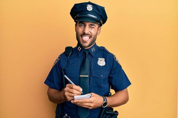 Handsome hispanic man wearing police uniform writing traffic fine sticking tongue out happy with funny expression. 