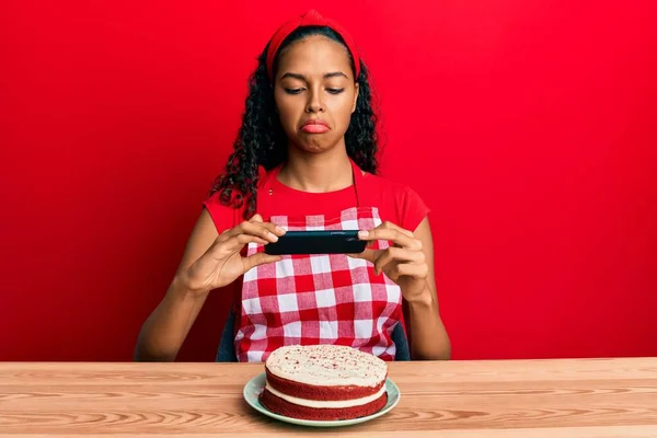 パン屋のエプロンを身に着けている若いアフリカ系アメリカ人の女の子は ニンジンケーキの写真を落ち込んで 苦痛を心配し 怒って恐れています 悲しい表情 — ストック写真