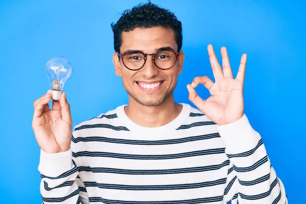 若いですハンサムなヒスパニック系の男は インスピレーションとアイデアのために電球を保持指でOkサインを行います 笑顔優しいジェスチャー優れたシンボル — ストック写真