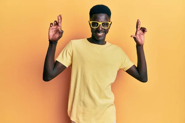年轻的非洲裔美国人 身穿休闲装 戴着眼镜 摆弄着手指 带着希望微笑 闭上眼睛 幸运与迷信的概念 — 图库照片