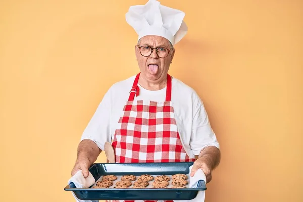 Starší Šedovlasý Muž Pekařské Uniformě Drží Domácí Sušenky Vystrkující Jazyk — Stock fotografie