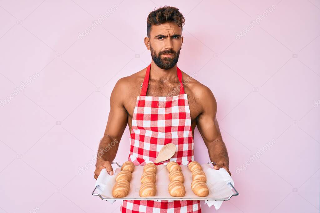 Young hispanic man shirtless wearing baker uniform holding homemade bread skeptic and nervous, frowning upset because of problem. negative person. 